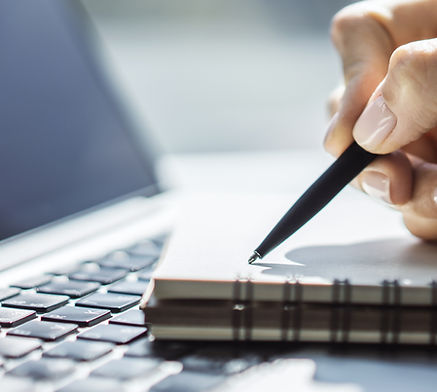 A hand, writing with a black pen in a spiral notebook which is resting on an open silver laptop.