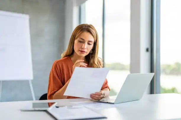 Woman reviewing a document at her desk while working on a laptop in a bright office