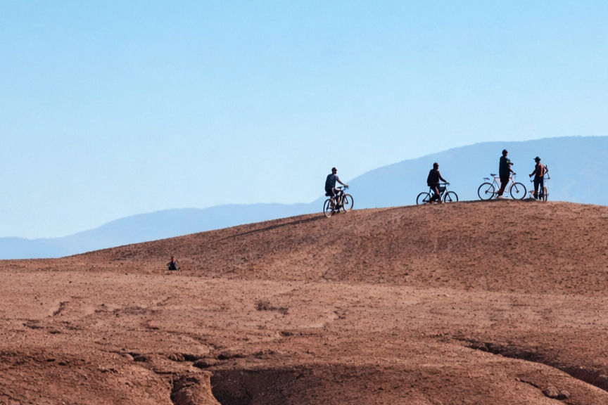 Cyclistes sur une colline désertique ocre, sous un ciel bleu, avec des montagnes floues en arrière-plan.