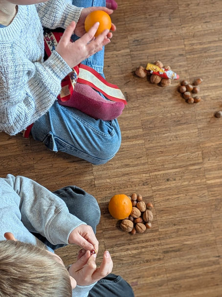 Kinder, die auf dem Boden sitzen und vor ihnen Nüsse undMandarinen liegen.