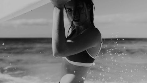 Model wearing minimalist swimwear holding a surfboard at the shoreline, photographed in natural coastal light.