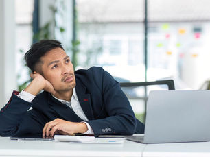 an asian man sitting at a desk with a laptop staring off into space