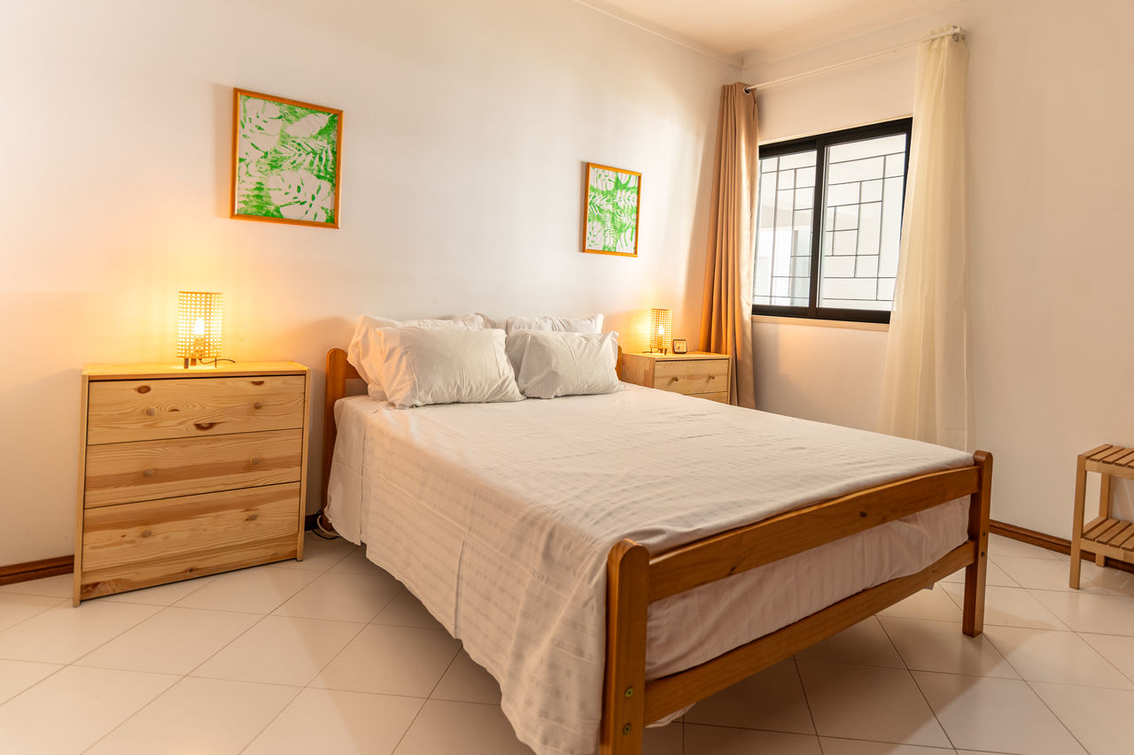 Bright bedroom with wooden bed, white bedding, bedside tables, and soft lighting, featuring a window with natural light.