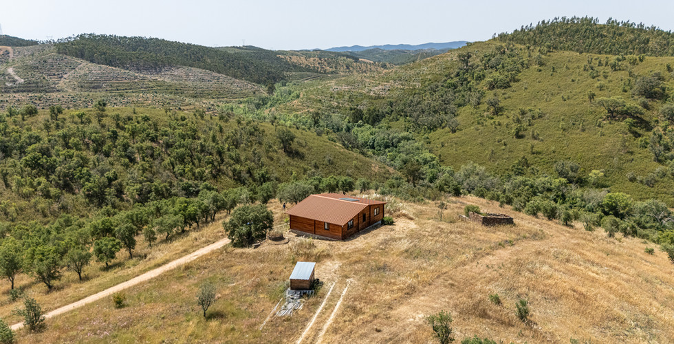 Aerial drone view of an off-grid wooden house near Sabóia, Alentejo, with access track and surrounding rural hills.