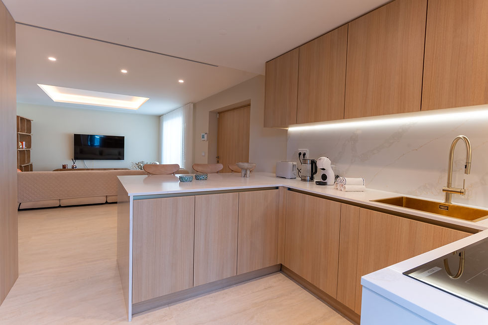 Cozinha moderna de conceito aberto com armários de madeira clara, bancadas brancas, pia dourada e sala de estar ao fundo.