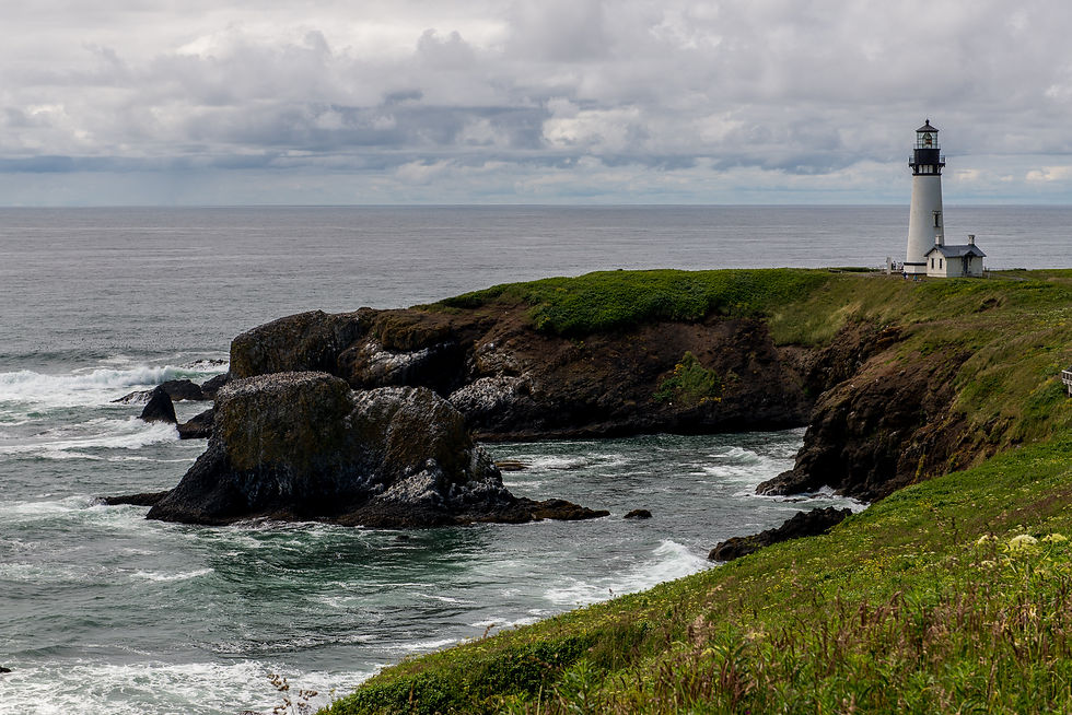 A white lighthouse stands on a grassy headland above dark coastal rocks and grey ocean surf.