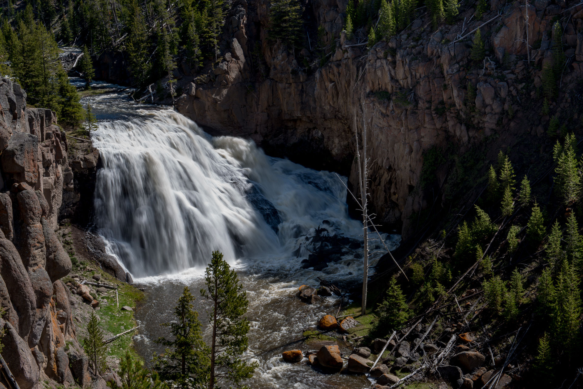 A broad white waterfall plunges over a rocky ledge into a gorge surrounded by conifers.