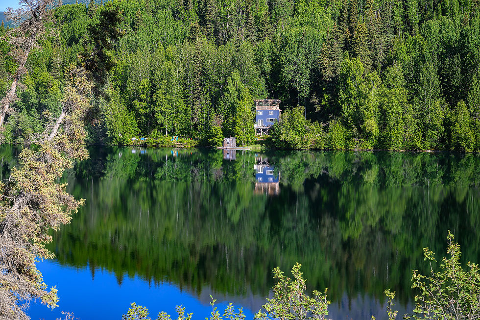 A small house stands at the edge of a densely forested lake, reflected with the trees in dark, still water.