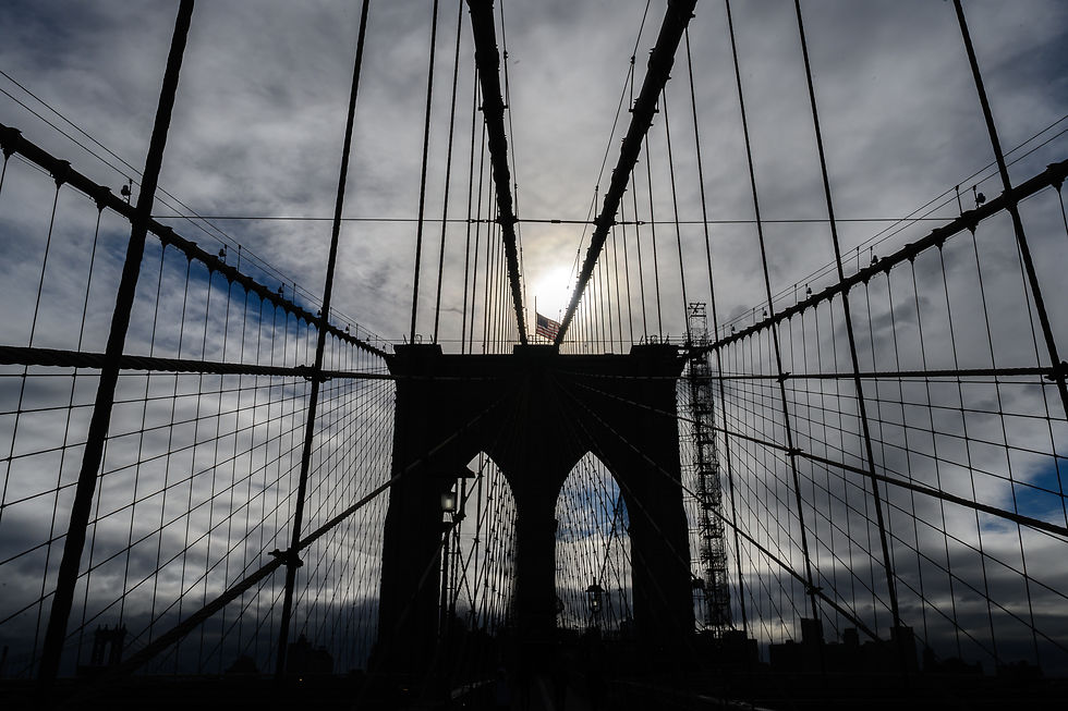 The stone arches and converging suspension cables of Brooklyn Bridge form a dark silhouette against sunlit clouds.