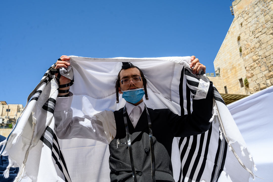 A young man lifts a white prayer shawl with black stripes around his shoulders beneath a clear blue sky.