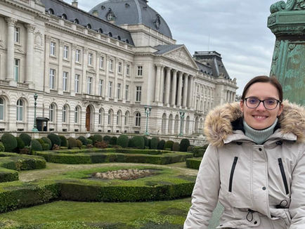 Téli városkép Brüsszelben, előtérben bundás kabátot viselő, szemüveges nő látható, mögötte a Királyi Palota klasszicista épülete, nyírt bokrokkal és szimmetrikus kerttel, borús időben.