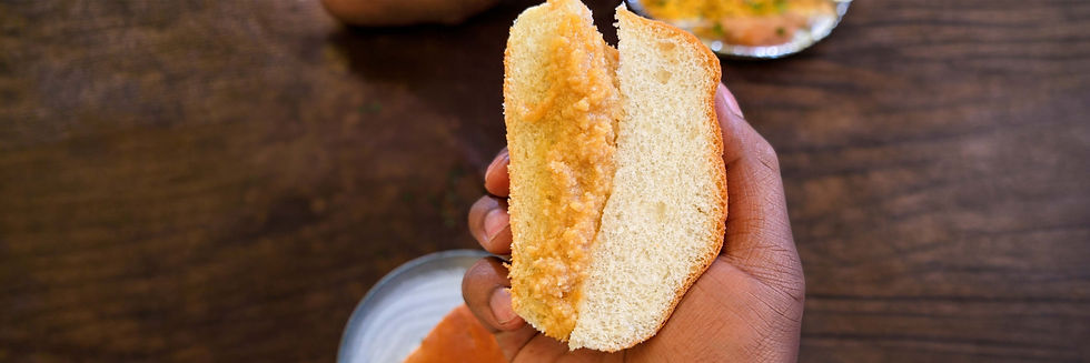 Hand holding split bun with kova filling, over wooden table. Plate with food visible in background. Warm tones.