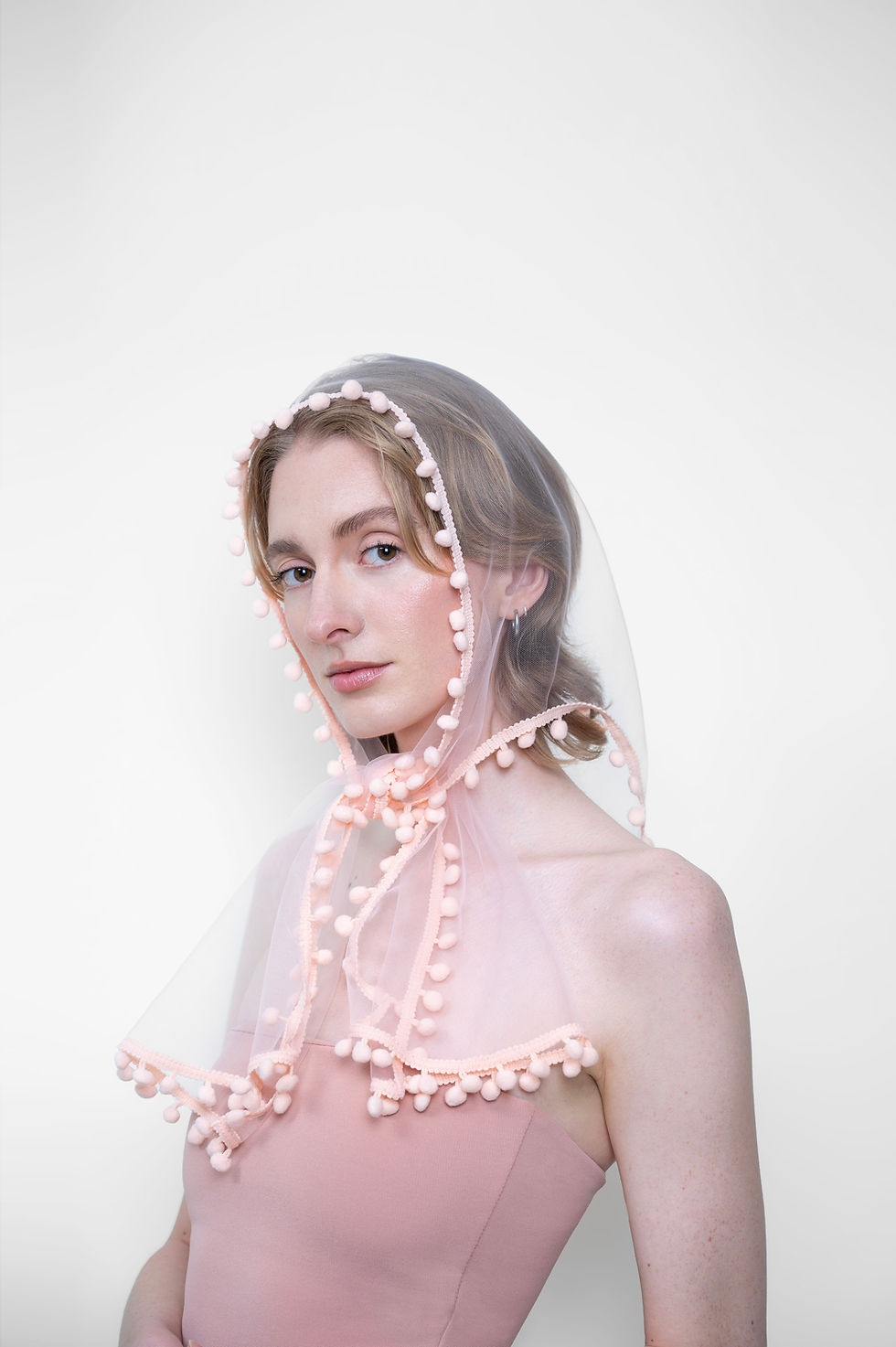 Person in a pink strapless dress and sheer hood with pom-poms, against a plain white background, gazing softly at the camera.