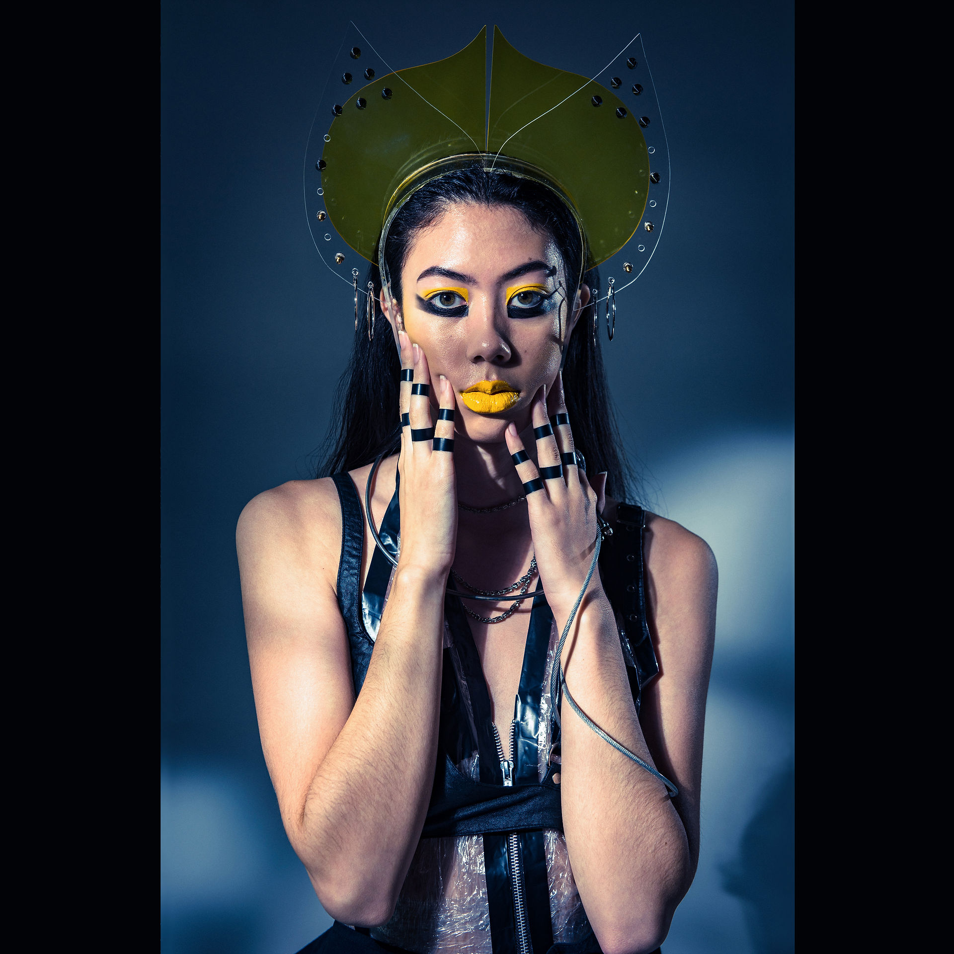 Fashion cyberpunk editorial with female model framing her face with her hands, her makeup is yellow and black graphic eye makeup, yellow lipstick, and she is wearing a yellow studded acrylic crown, plastic halter top, leather half-bra, metal wires and chains adorn her neck and arm, and her fingers are taped with black PVC tape..