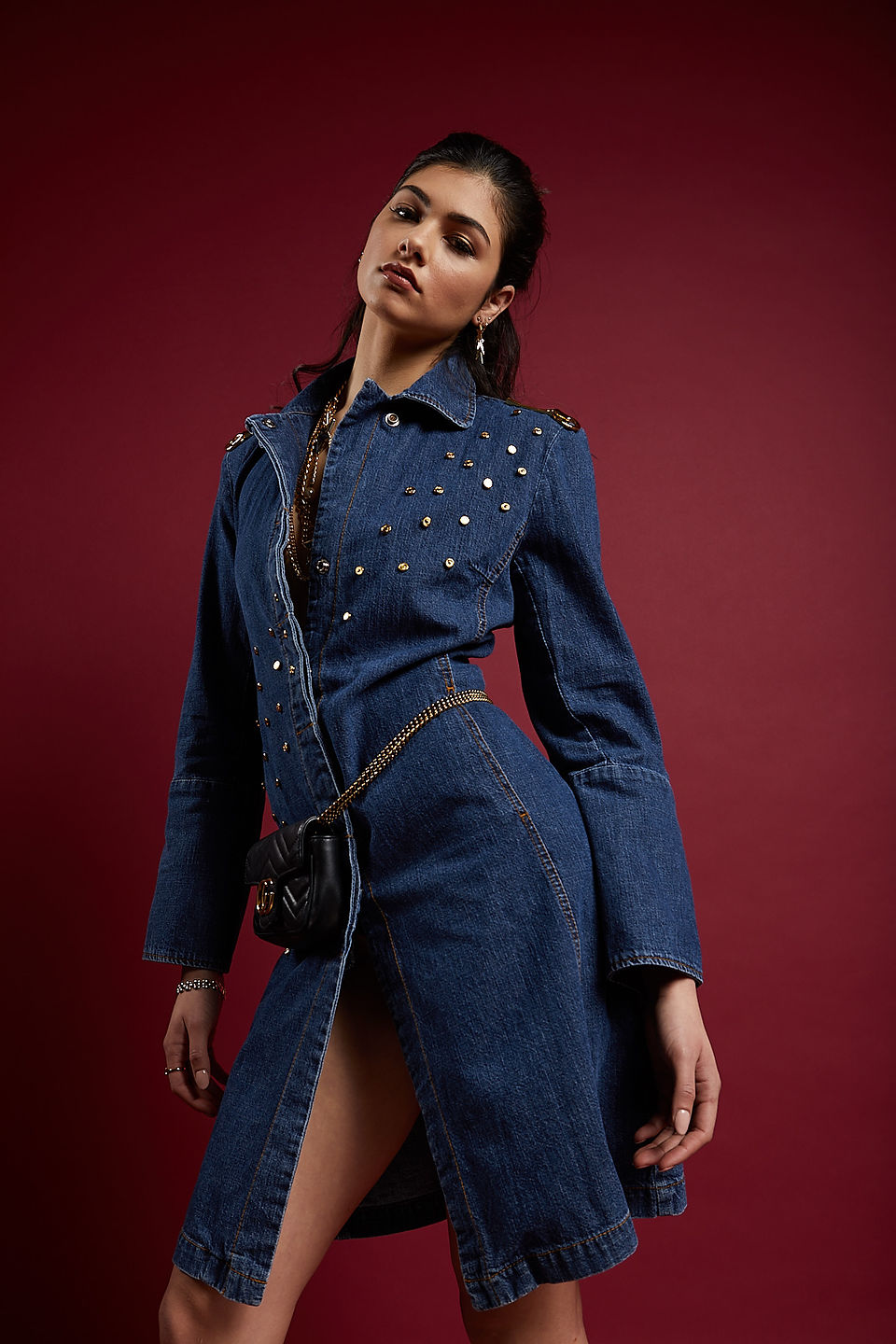Female model standing in a S-shape pose wearing a denim dress studded with gold hardward and accessorized with gold chain necklaces and a black leather with gold chain Gucci hip purse