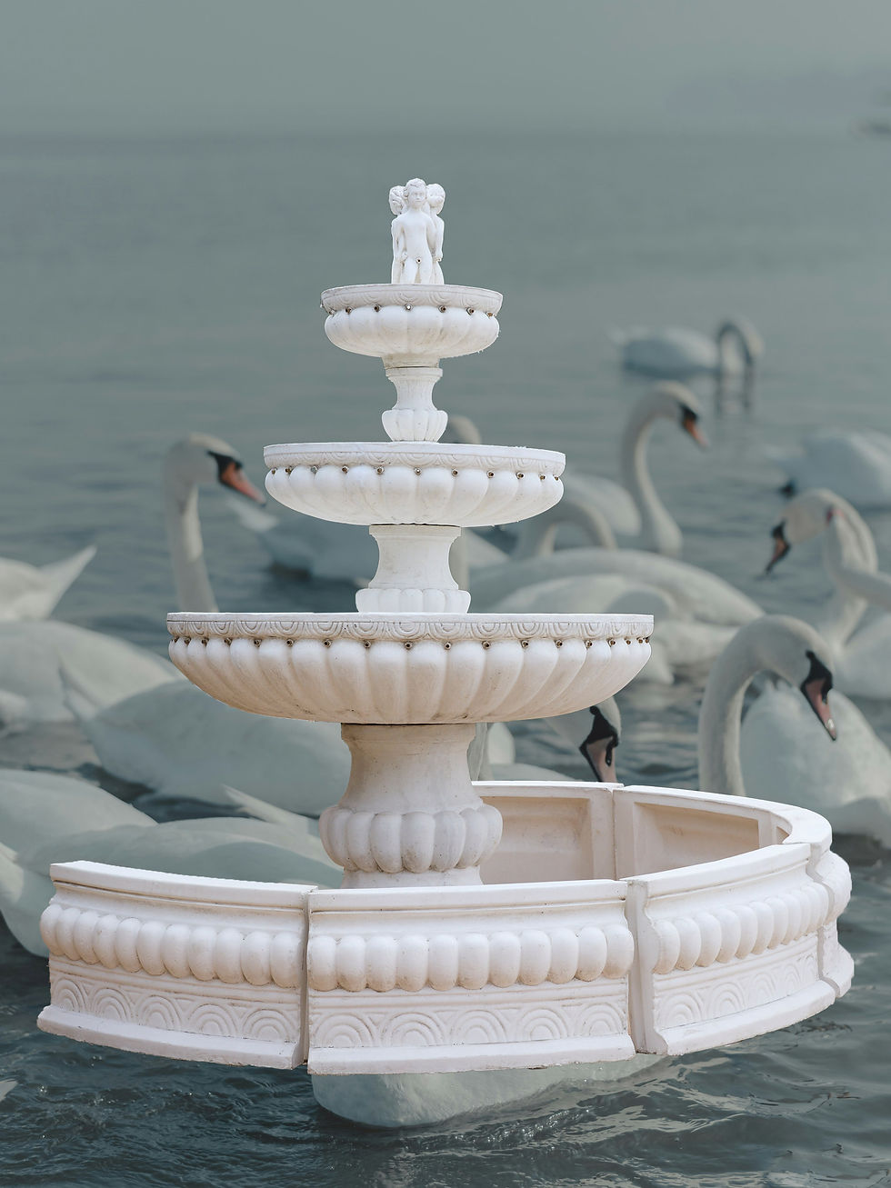 White stone fountain with three tiers and a figure of three children at the top, surrounded by white swans in a body of water
