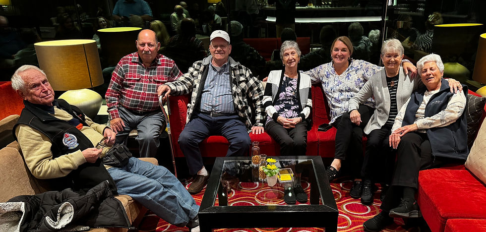 Seven smiling people sit on red sofas in a warmly lit room. A glass table holds flowers and snacks, with reflections visible in the background.