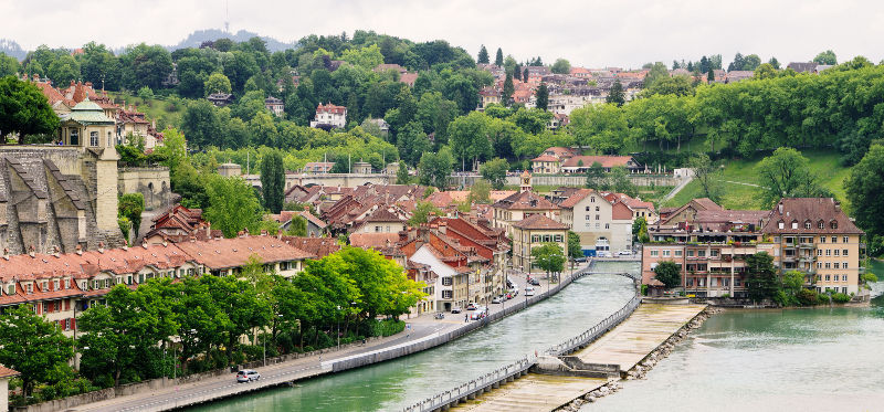 A picturesque view of Verona, nestled along a river, featuring charming red-roofed buildings surrounded by lush green hills.