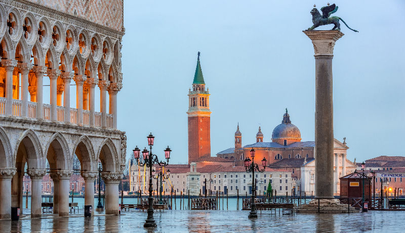 Morning light illuminates the iconic architecture of St. Mark's Square in Venice, highlighting the Doge's Palace, the Campanile, and historic sculptures overlooking the tranquil water.
