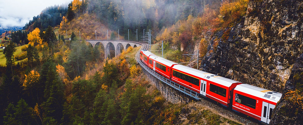 Red train crosses a stone viaduct in a misty, autumnal forest with vibrant yellow and green foliage on surrounding hills.