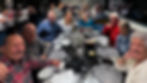 Group of six sitting around a restaurant table, smiling. A server behind them makes a peace sign. Warm atmosphere, patterned placemats.