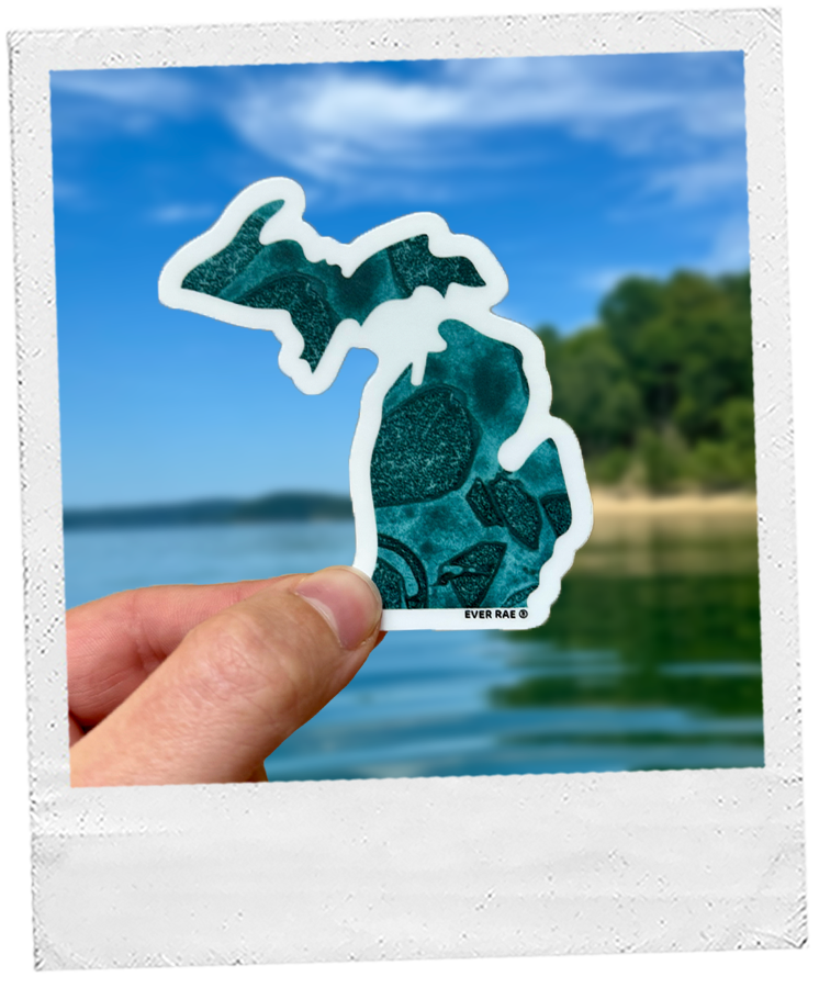 Thumbnail: Hand holding a Michigan-shaped sticker in a deep teal color, with a calm lake and tree-lined shore