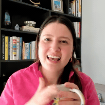 Taylor Harrington speaking enthusiastically on video call in bright pink shirt, holding a coffee mug, with bookshelf visible behind her.