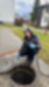 Robert Novogradec in grauer Arbeitskleidung mit Kapuze und blauen Einweghandschuhen kniet neben einem geöffneten Kanaldeckel und hält einen Hochdruckschlauch in die Kanalöffnung. Wasser läuft aus der Düse des Schlauchs. Er lächelt in die Kamera und zeigt einen Daumen hoch. Im Hintergrund sind gepflegte Grünflächen, Wohnhäuser und schneebedeckte Berge unter einer bewölkten Himmel zu sehen.