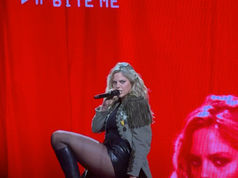 Performer kneels on stage with a microphone, wearing a fringed jacket and black boots. Red background displays "BITE ME" text.