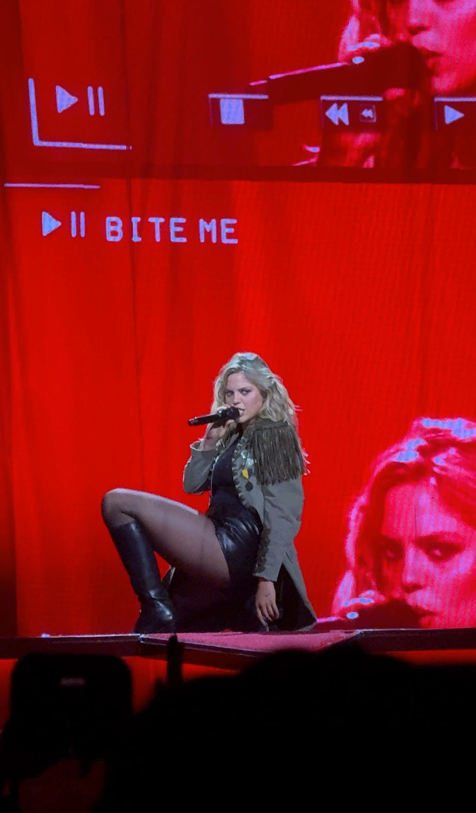 Performer kneels on stage with a microphone, wearing a fringed jacket and black boots. Red background displays "BITE ME" text.