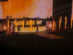 Rock band performs on a multi-level stage with flames, orange lighting, and a drummer at right in a dramatic concert scene