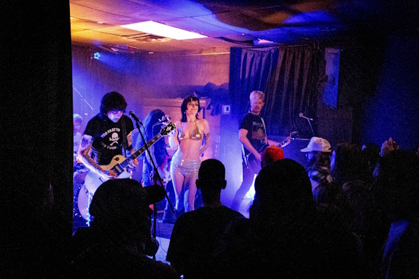 Band performing on stage with lead singer and guitarists. Dimly lit venue with blue and orange lights. Audience silhouetted in foreground.