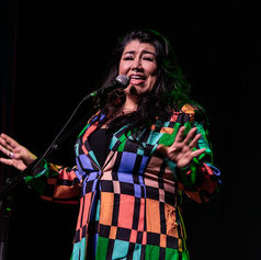 Singer performs on a dark stage at a microphone, wearing a colorful checkered dress and gesturing expressively.