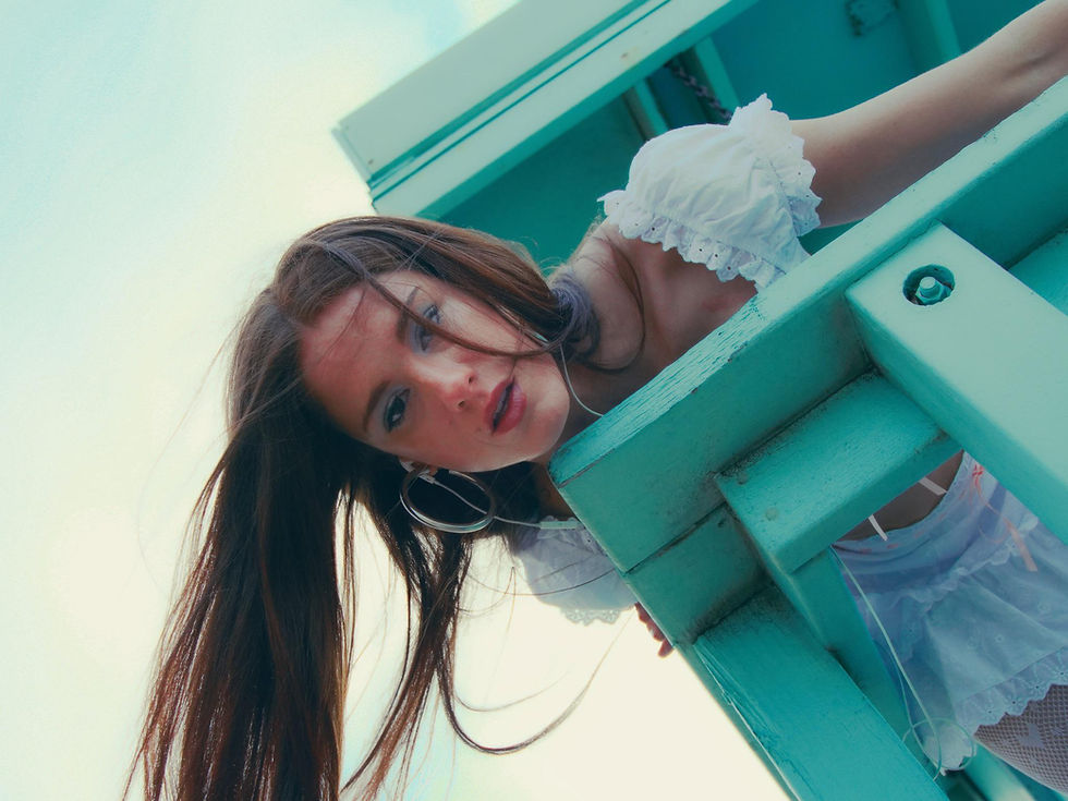 Woman in a white dress leans over a turquoise railing, gazing down with long hair blowing. Sky is overcast, creating a serene mood.