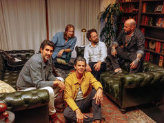 Five men (band of heathens) gather in a cozy room with leather sofas and bookshelves. A vintage rug and chandelier add warmth. Mood is relaxed and sociable.