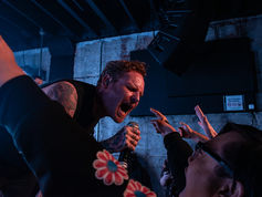 Singer passionately performs to an enthusiastic crowd in a dimly lit venue. Fans reach out; one wears a sweatshirt with colourful flowers.