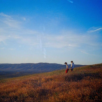 Two hikers walk across a grassy hillside under a clear blue sky, with distant mountains and a calm, open landscape.