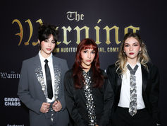 The three sisters that make up the rock trio The Warning in embellished suits stand against a black backdrop with "The Warning" text. They have serious expressions and styled hair.