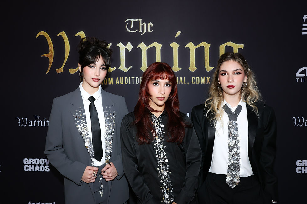 The three sisters that make up the rock trio The Warning in embellished suits stand against a black backdrop with "The Warning" text. They have serious expressions and styled hair.