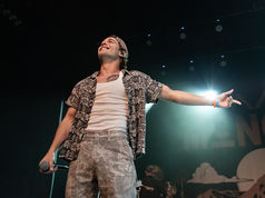 Singer on stage, smiling with arms outstretched, holding a mic. Wears patterned shirt, backward cap. Bright lights and partial background text.