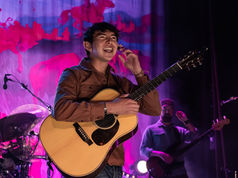 Musician with guitar smiles on stage, vibrant backdrop with red and purple hues. Another performer holds a bass guitar in the dim background.