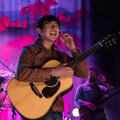 Musician with guitar smiles on stage, vibrant backdrop with red and purple hues. Another performer holds a bass guitar in the dim background.