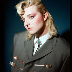 Person with blond hair and pink makeup in a gray suit adorned with cutlery. Dark blue background adds a dramatic mood.