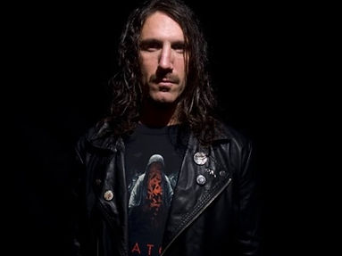 Johnny Stewart (Pillars of a Twisted City) with long hair and mustache in black leather jacket, graphic t-shirt with abstract art, against dark background, serious expression.