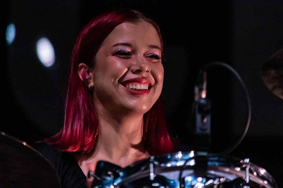 Smiling woman with red hair plays drums, exuding joy. Dark background, reflective drum surfaces.