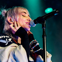 Singer with blonde hair performs passionately on stage, eyes closed, hands near face. Wears a light jacket. Blue-green stage lights illuminate.