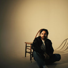 Bearded man in black jacket sits on the floor beside a wooden chair in a warm, minimalist studio, looking calm.