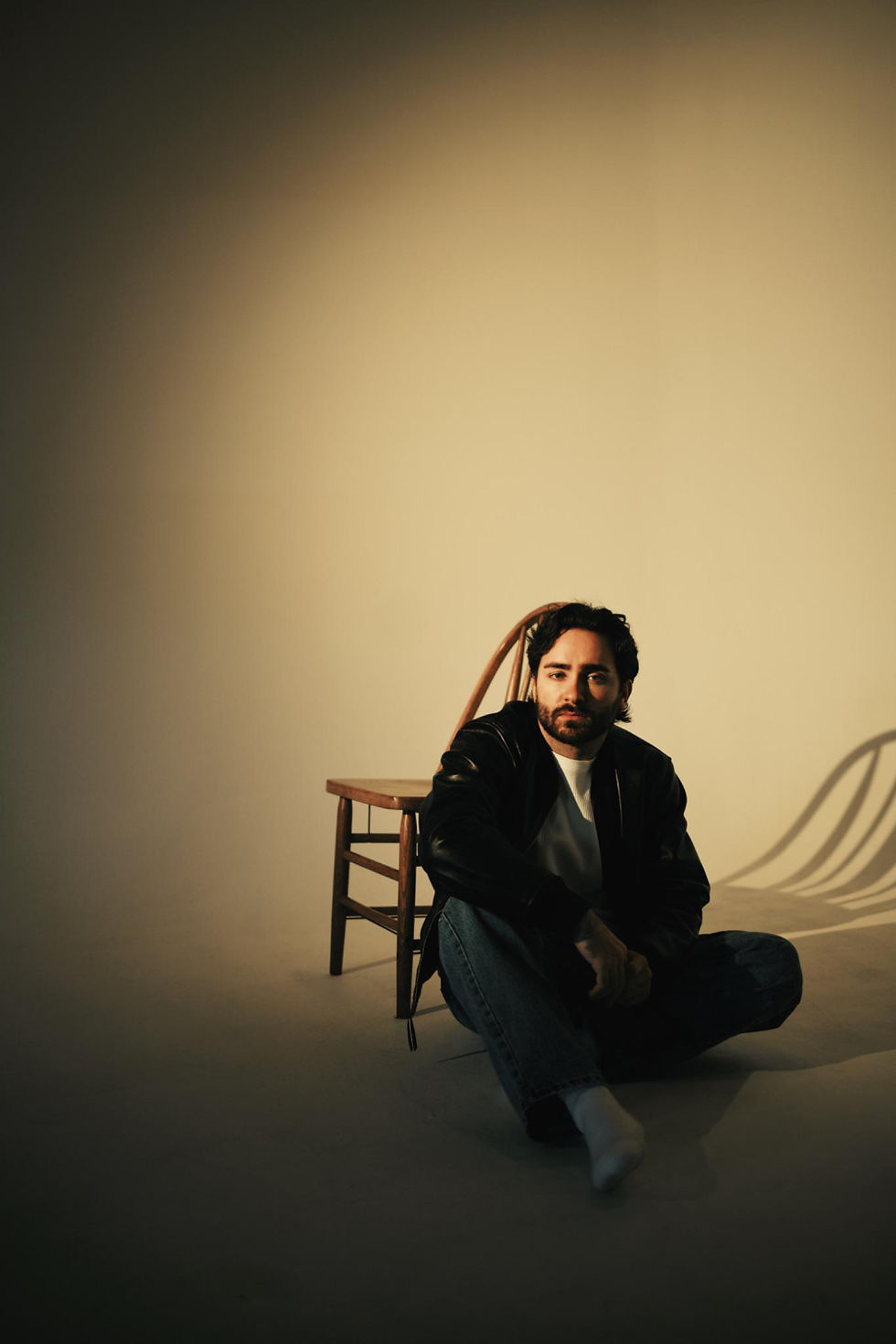 Bearded man in black jacket sits on the floor beside a wooden chair in a warm, minimalist studio, looking calm.