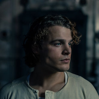 Young person (Max McNown) with curly hair gazes to the side in a dimly lit room. Wearing a light shirt, their expression appears contemplative. Dark background.