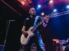 Guitarist plays passionately under purple stage lights with a drummer in the background. Dark curtains and a Yamaha drum set visible.